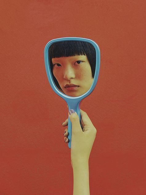 fotografia de uma mulher segurando um espelho retrô, de cabo azul, frente de uma parede cor de vinho. No reflexo do espelho, aparece o rosto de uma mulher chinesa, com a franja cortada bem reta.
