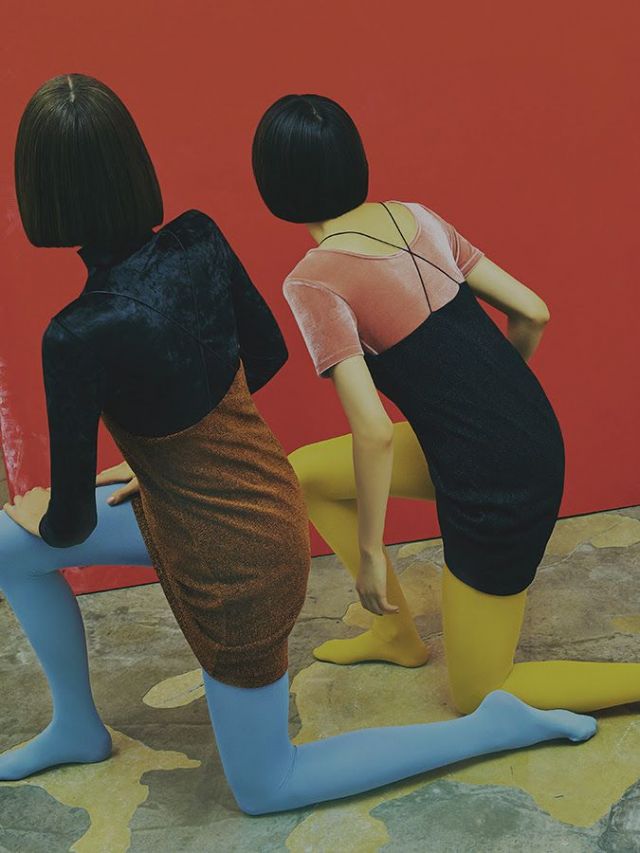 Fotografia de duas mulheres fazendo o mesmo movimento, apoiadas sobre um joelho só, meio agachadas, sendo vistas de costas. Usam o cabelo curto, preto e bem liso. Uma usa o vestido de camurça marrom sobre uma blusa de mangas longas preta e uma meia-calça azul pastel. A outra usa um vestido preto sobre uma blusa rosa e meia-calça amarela. Elas estão olhando para a mesma parede cor de vinho.