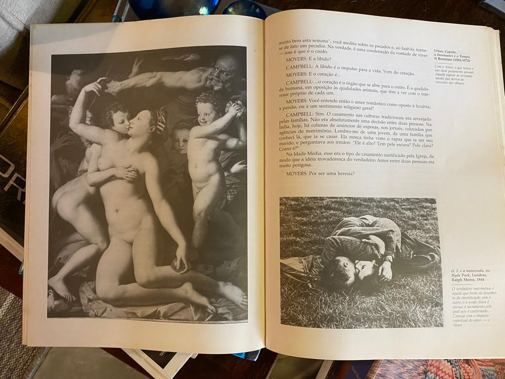 Foto da página aberta de um livro. Na página da esquerda, uma pintura clássica de uma mulher nua sendo beijada e acariciada por anjos. "Vênus, Cupido, a Insensatez e o Tempo". Na página direita, o texto: "MOYERS: E a libido? CAMPBELL: A libido é o impulso para a vida. Vem do coração. MOYERS: E o coração é... CAMPBELL: ... o coração é o órgão que se abre para o outro. É a qualidade humana, em oposição às qualidades animais, que têm a ver com o interesse próprio de cada um. MOYERS: Você entende então o amor romântico como oposto à luxúria, à paixão ou a um sentimento religioso geral? CAMPBELL: Sim. O casamento nas culturas tradicionais era arranjado pelas famílias. Não era absolutamente uma decisão de duas pessoas. Na Índia, hoje, há colunas de anúncios de esposas, nos jornais, colocados por agências de matrimônio. Lembro-me de uma jovem, de uma família que conheci lá, que ia se casar. Ela nunca tinha visto o rapaz que ia ser seu marido, e perguntava aos irmãos: "Ele é alto? Tem pele escura? Pele clara? Como é?" Na idade média, esse era o tipo de casamento santificado pela Igreja, de modo que a ideia trovadoresca do verdadeiro Amor entre duas pessoas era muito perigosa. MOYERS: Por ser uma heresia?" Abaixo, uma foto de um homem e uma mulher abraçados e deitados na grama. "G.I. e a namorada, no Hyde Park. Londres, Ralph Morse, 1944".
