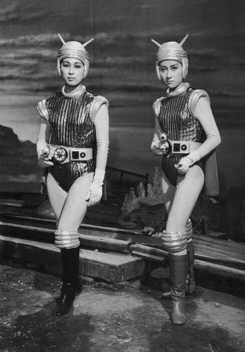Fotografia antiga, em preto e branco, de duas mulheres vestidas com collants espaciais, cinturão cheio de botões, capa e capacetes de anteninhas. Elas apontam armas lasers para a câmera, uma delas com bastante cara de brava.